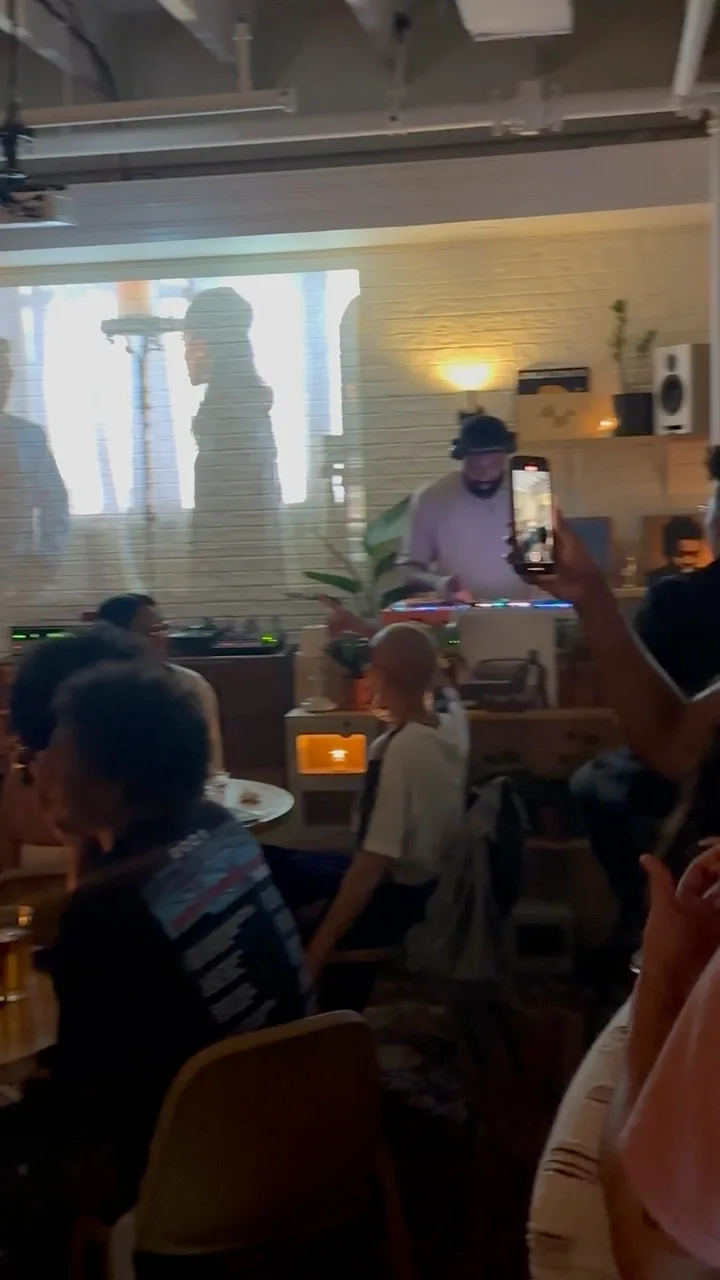 People gathered around small tables in a warmly lit lounge, with a DJ setup and projected imagery in the room