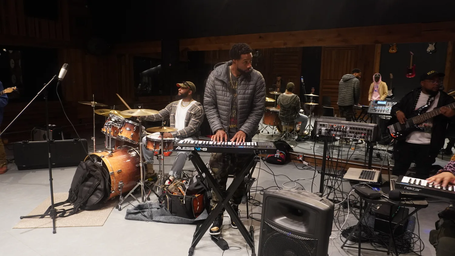 Musicians gathered around keyboards, drums, and bass in a rehearsal room